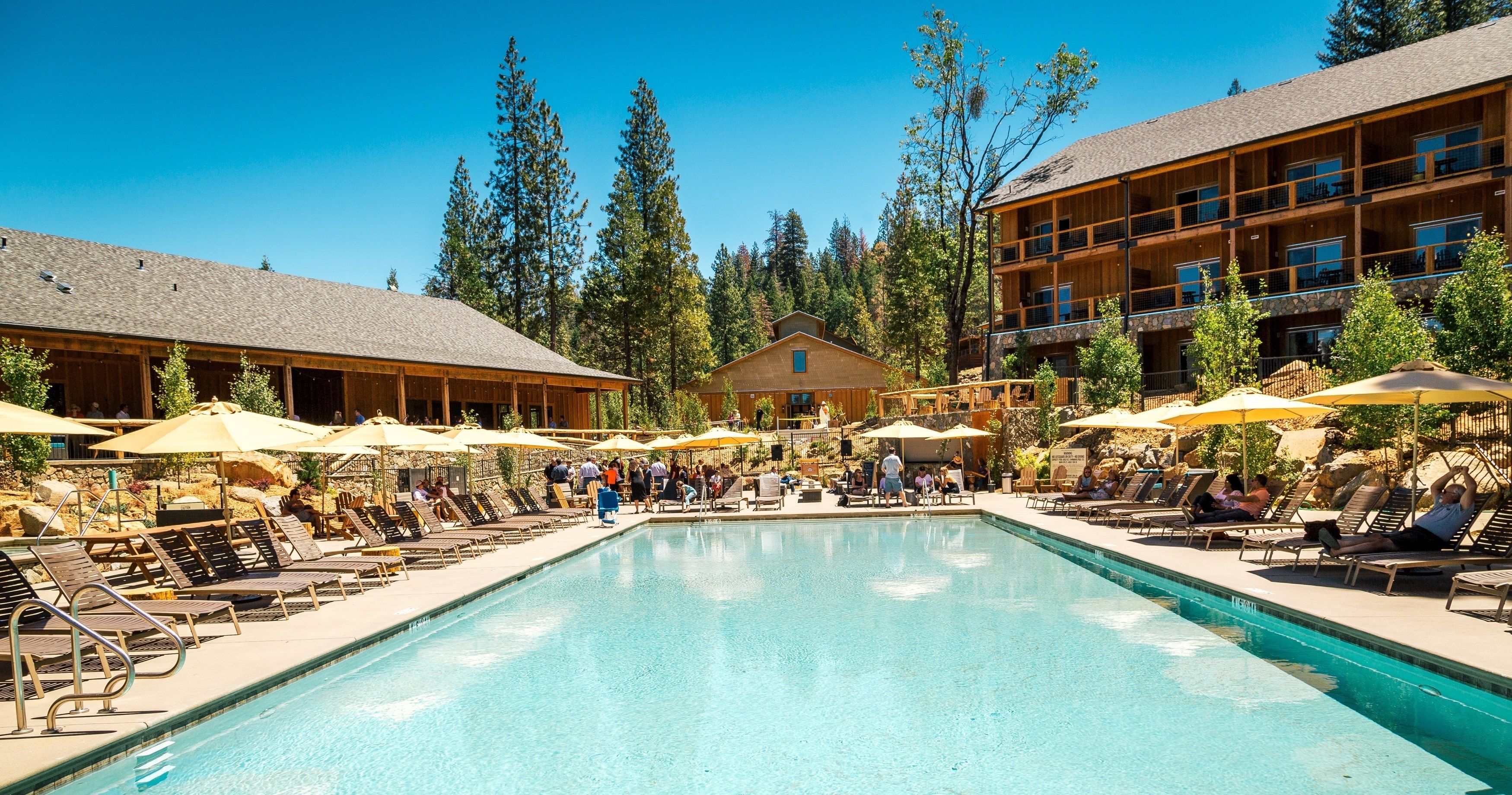 A pool area with lounge chairs and umbrellas, surrounded by wooden buildings and trees.