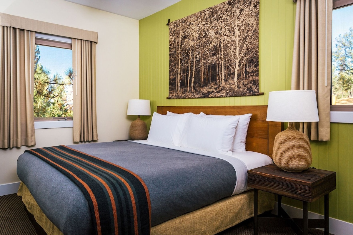 A hotel room with a bed, two bedside lamps, a window with curtains, and a wall painting.