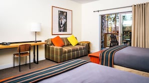 A hotel room with a desk, a plaid sofa with orange and yellow cushions, a bed with striped bedding, and a window with curtains.
