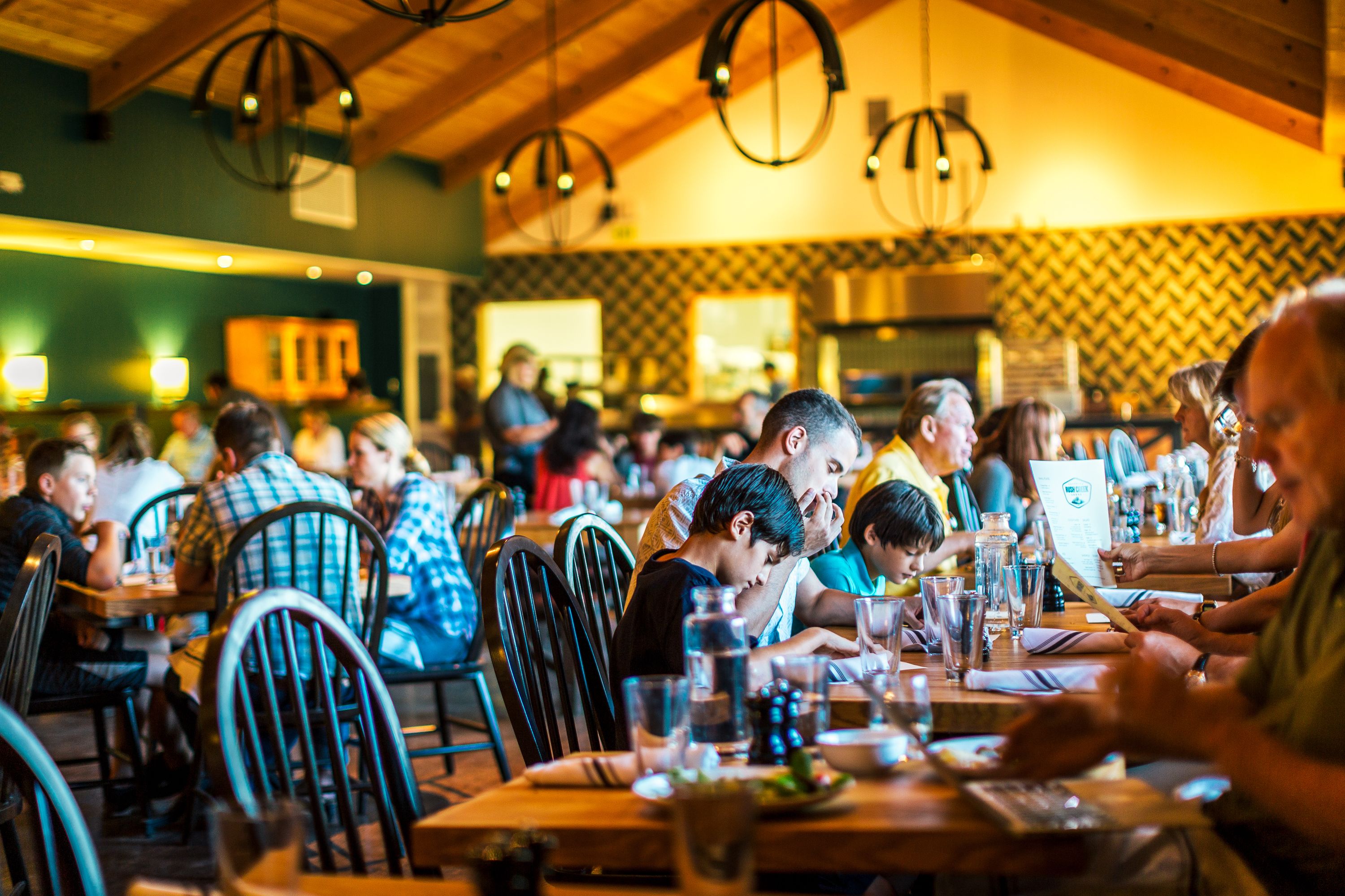 A restaurant with wooden tables and chairs, people dining, and hanging pendant lights.