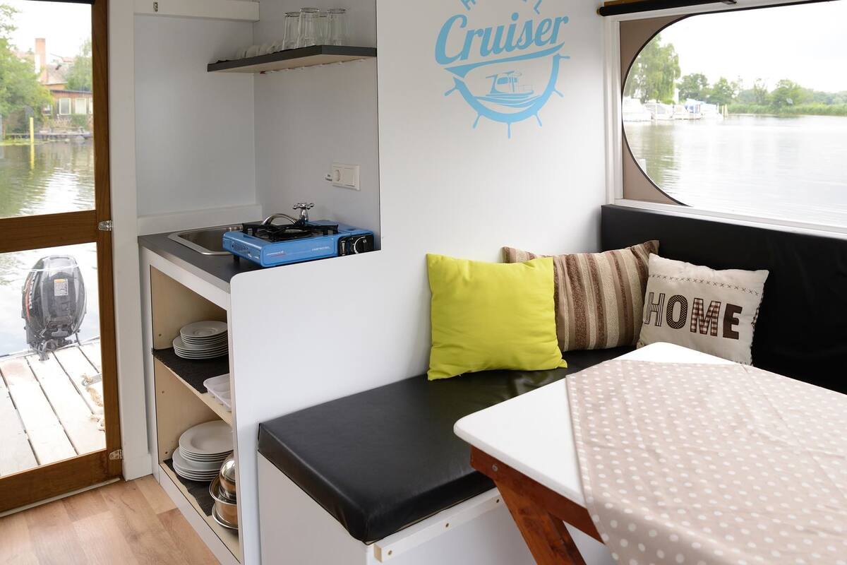 A compact kitchen with a blue portable stove, a sink, and a wall-mounted shelf with glass jars. A bench with pillows and a "HOME" pillow is visible.