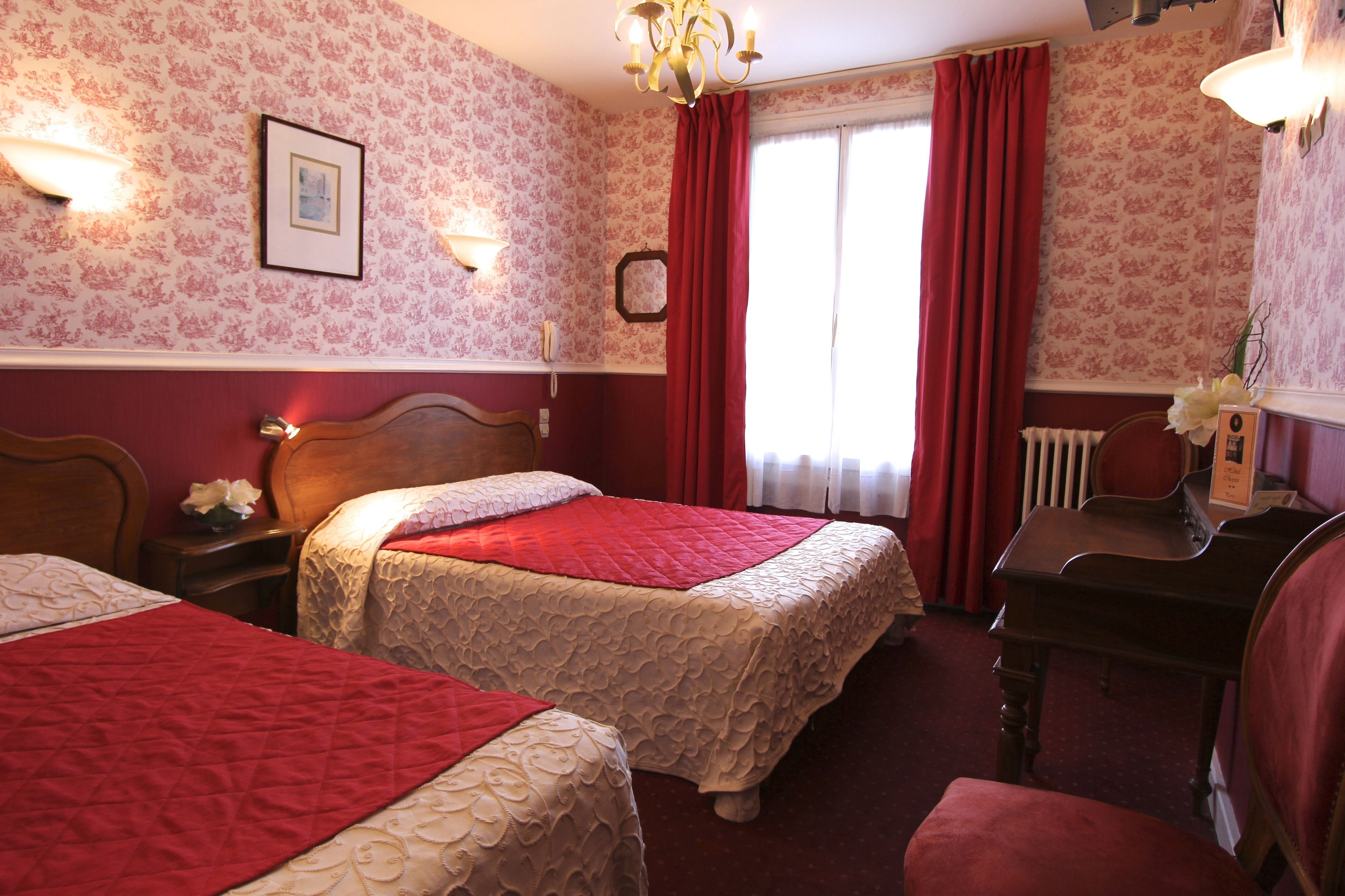 A hotel room with two beds, a wooden desk, a radiator, and a window with red curtains.
