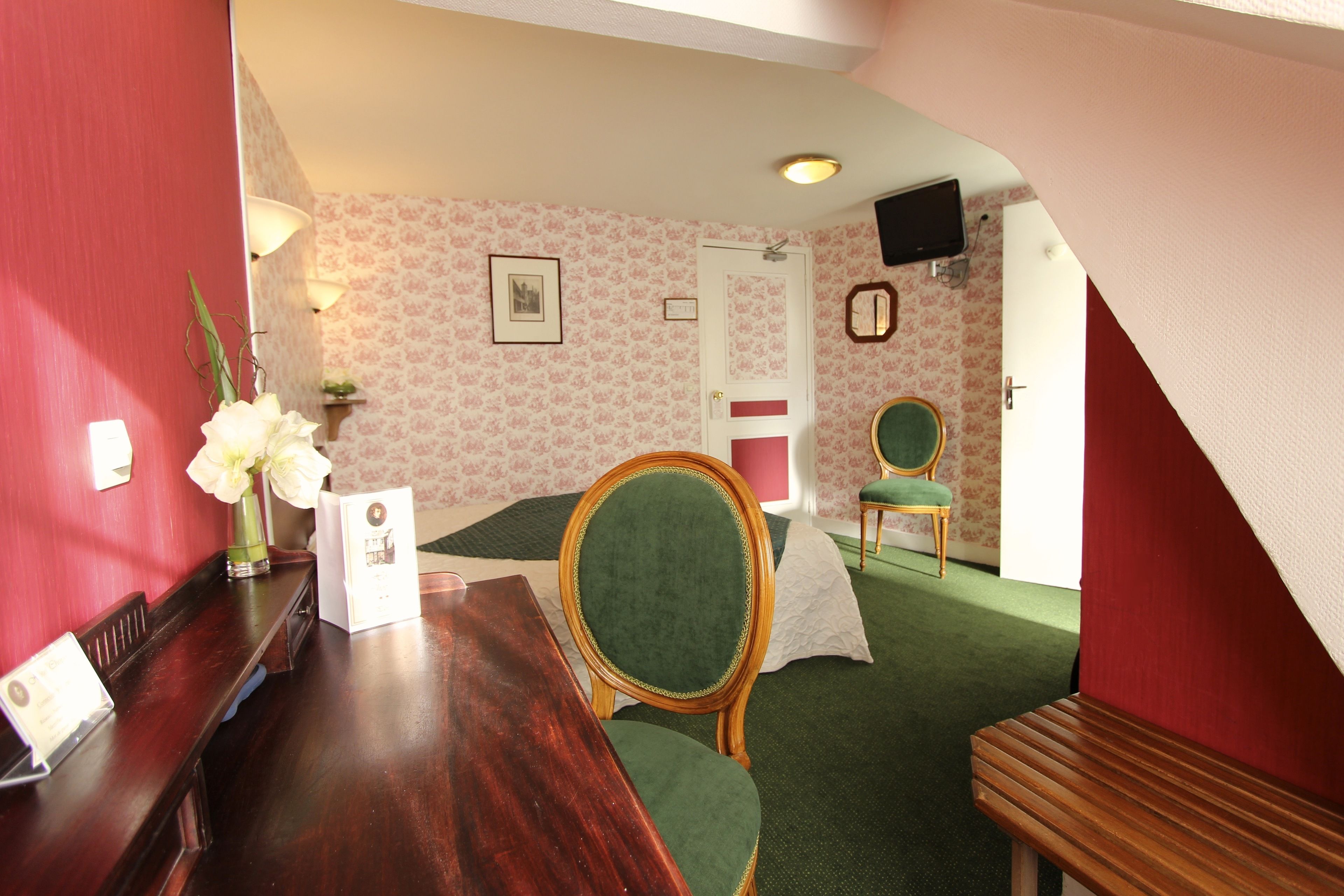 A room with a wooden desk, a green upholstered chair, a white door, and a television mounted on the wall.