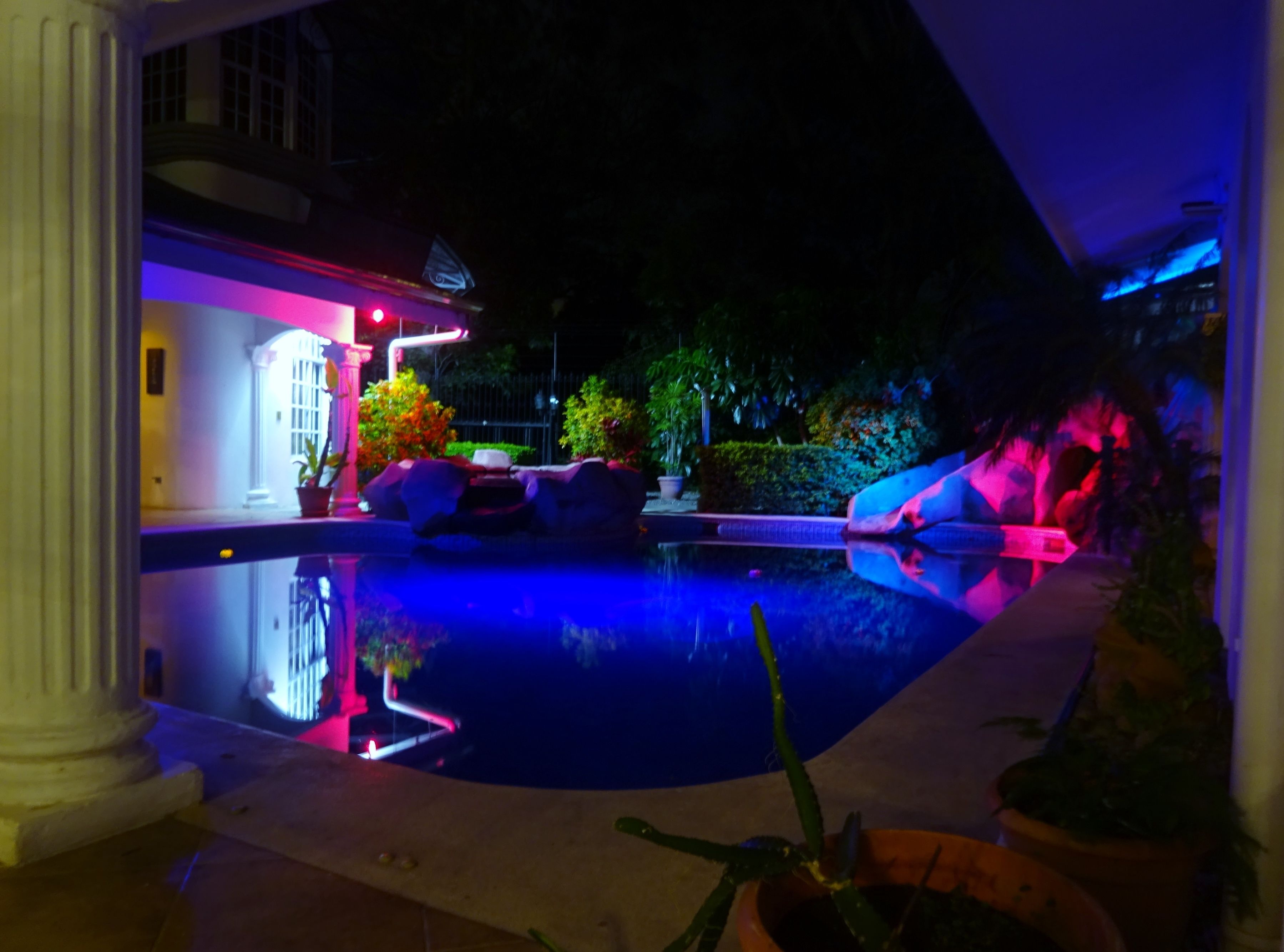 A nighttime view of a swimming pool with a patio area, surrounded by potted plants and a building with illuminated windows.