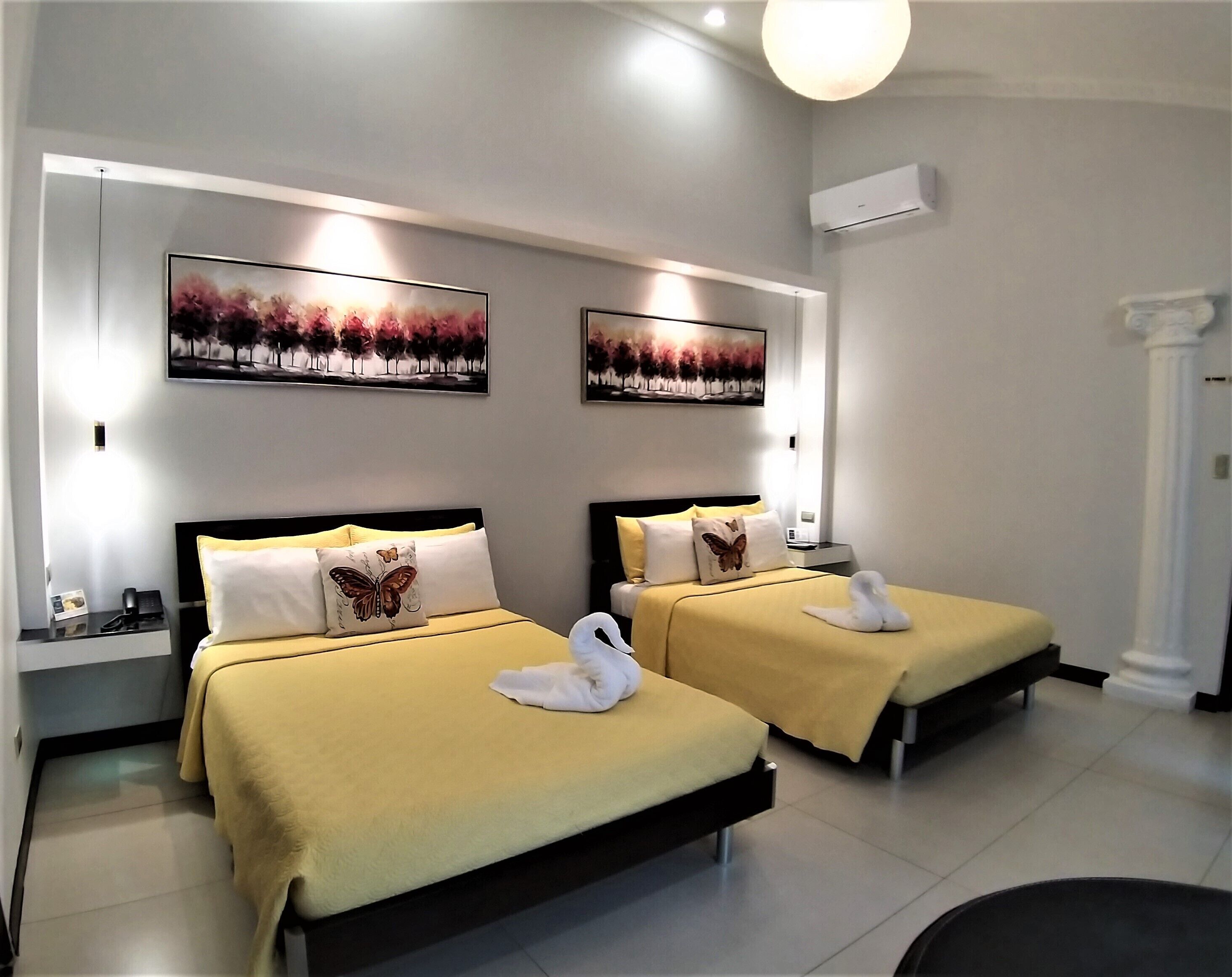 Habitación de hotel con dos camas, cada una con una colcha amarilla y una decoración de cisne blanco. Se observan dos unidades de aire acondicionado fijadas a la pared y dos cuadros enmarcados sobre las camas.