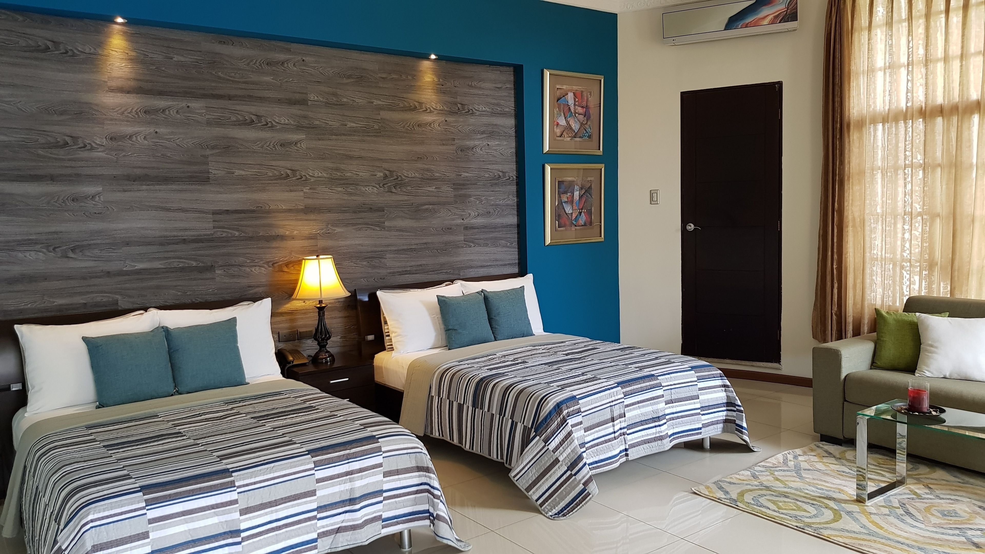 A hotel room with two beds, a nightstand, a lamp, a sofa, and a door.