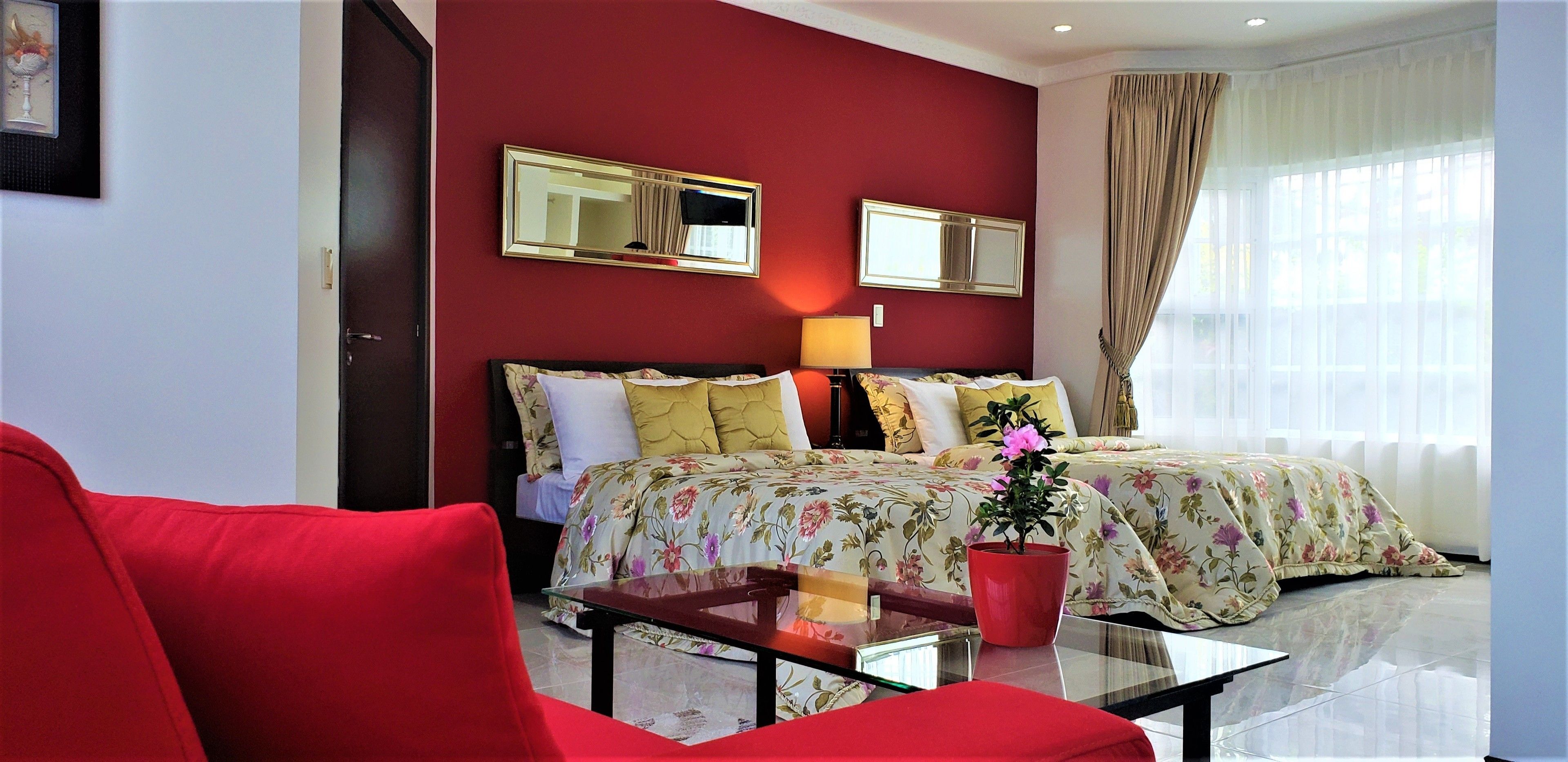 A hotel room with a red wall, two beds with floral bedding, a red armchair, a glass coffee table, and a potted plant.