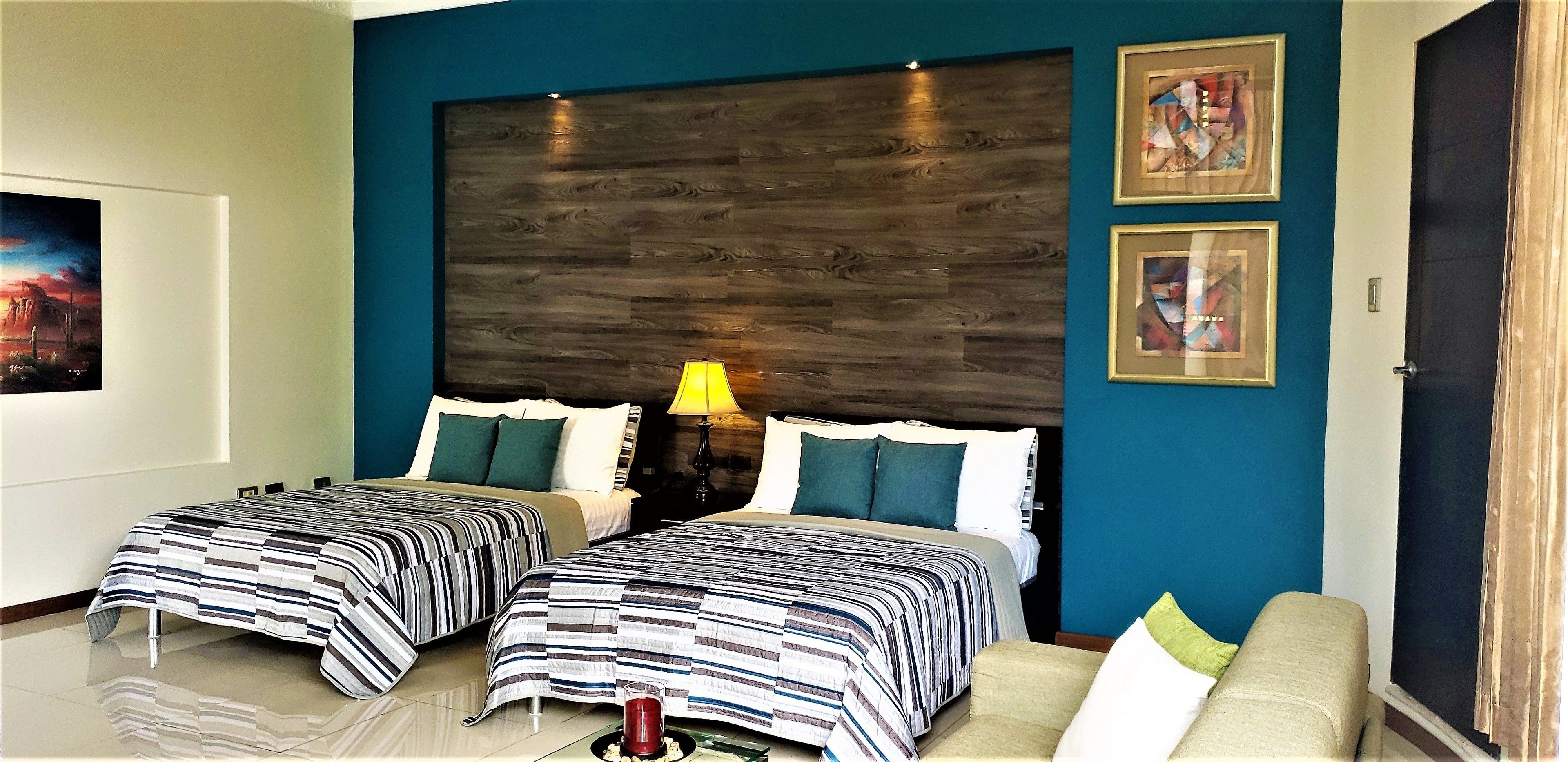 A hotel room with two beds, a nightstand with a lamp, a sofa, and artwork on the walls.