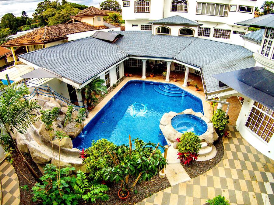 A large, rectangular swimming pool with a stone border and a small hot tub, surrounded by potted plants and a checkered tile deck.