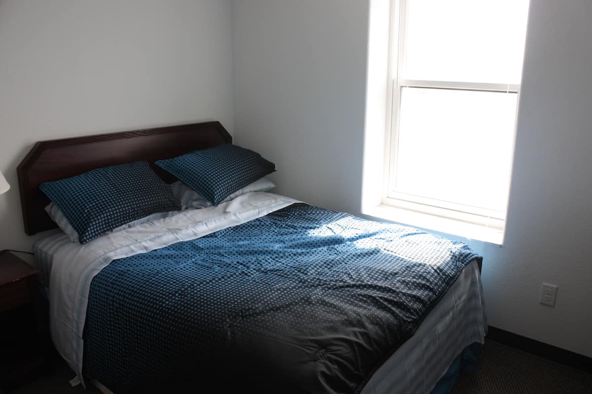 A neatly made bed with a blue and white patterned bedspread and pillows, a wooden headboard, and a window providing natural light.