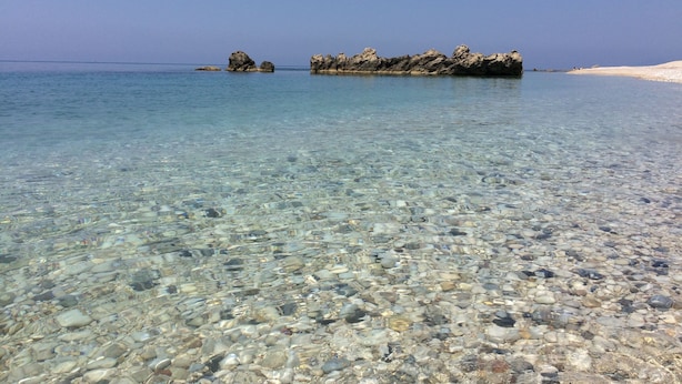Uma praia clara com águas rasas e pedras, um afloramento rochoso e uma área arenosa.