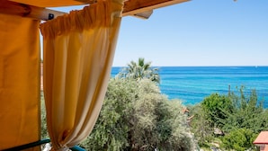 Vista de varanda com pérgola de madeira e cortinas amarelas, com paisagem costeira, árvores e o mar.