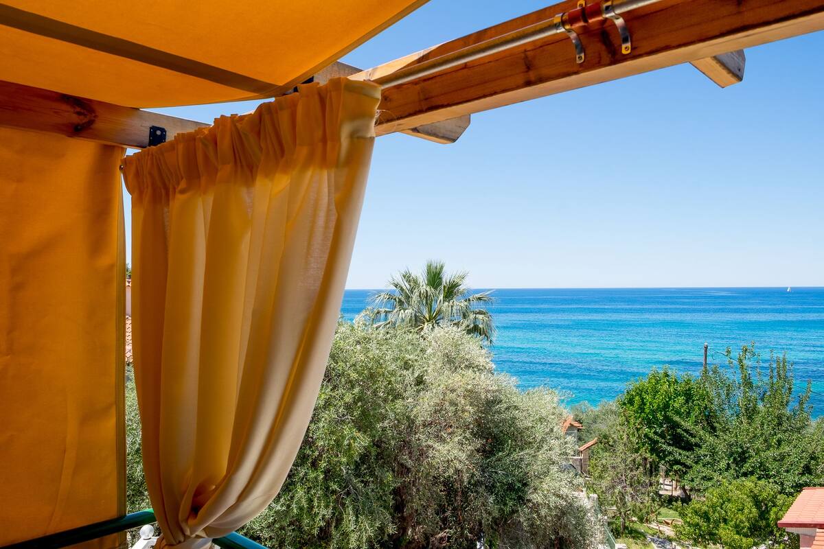 A view from a balcony with a wooden pergola and yellow curtains, overlooking a coastal landscape with trees and the sea.
