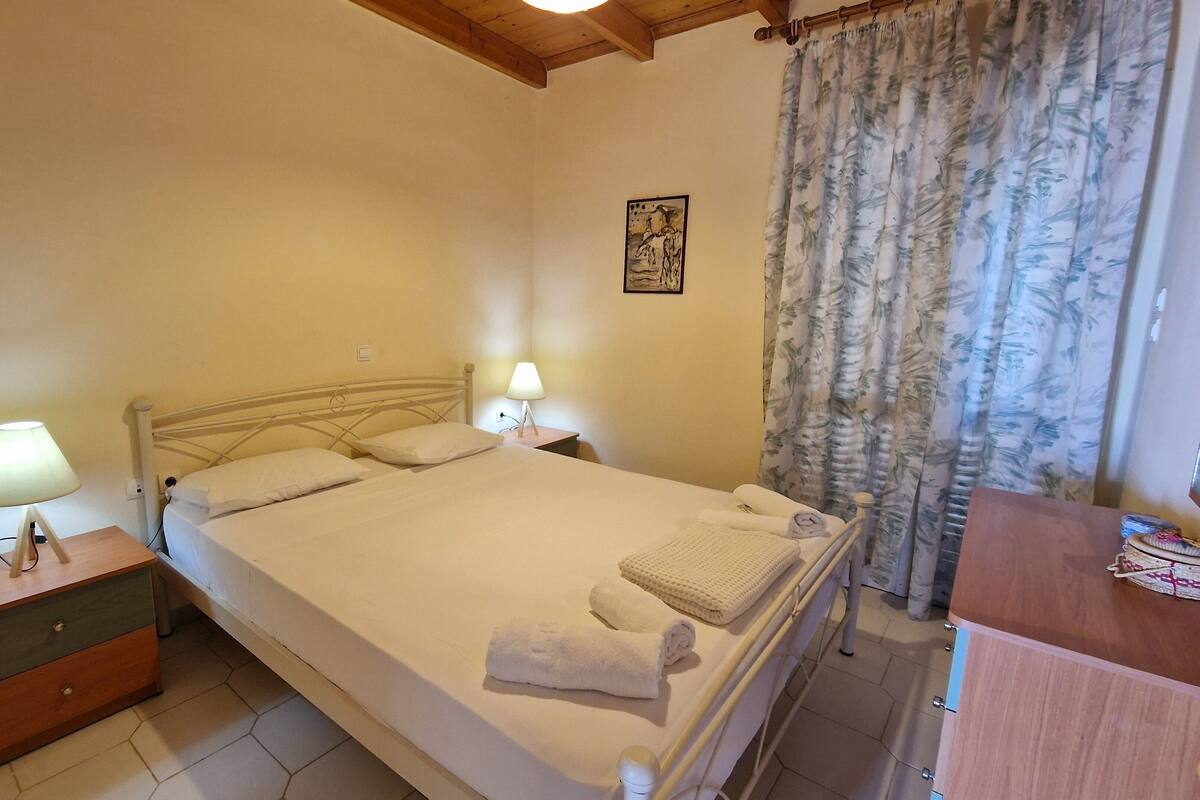 A neatly made bed with white linens and two bedside lamps, a wooden nightstand, a wooden dresser, and a floral curtain.