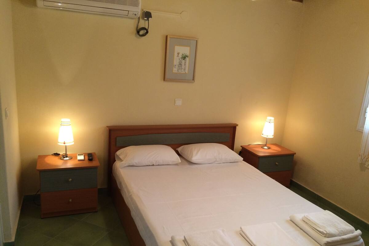 A hotel room with a single bed, two bedside tables, a wall-mounted air conditioner, and a framed picture on the wall.