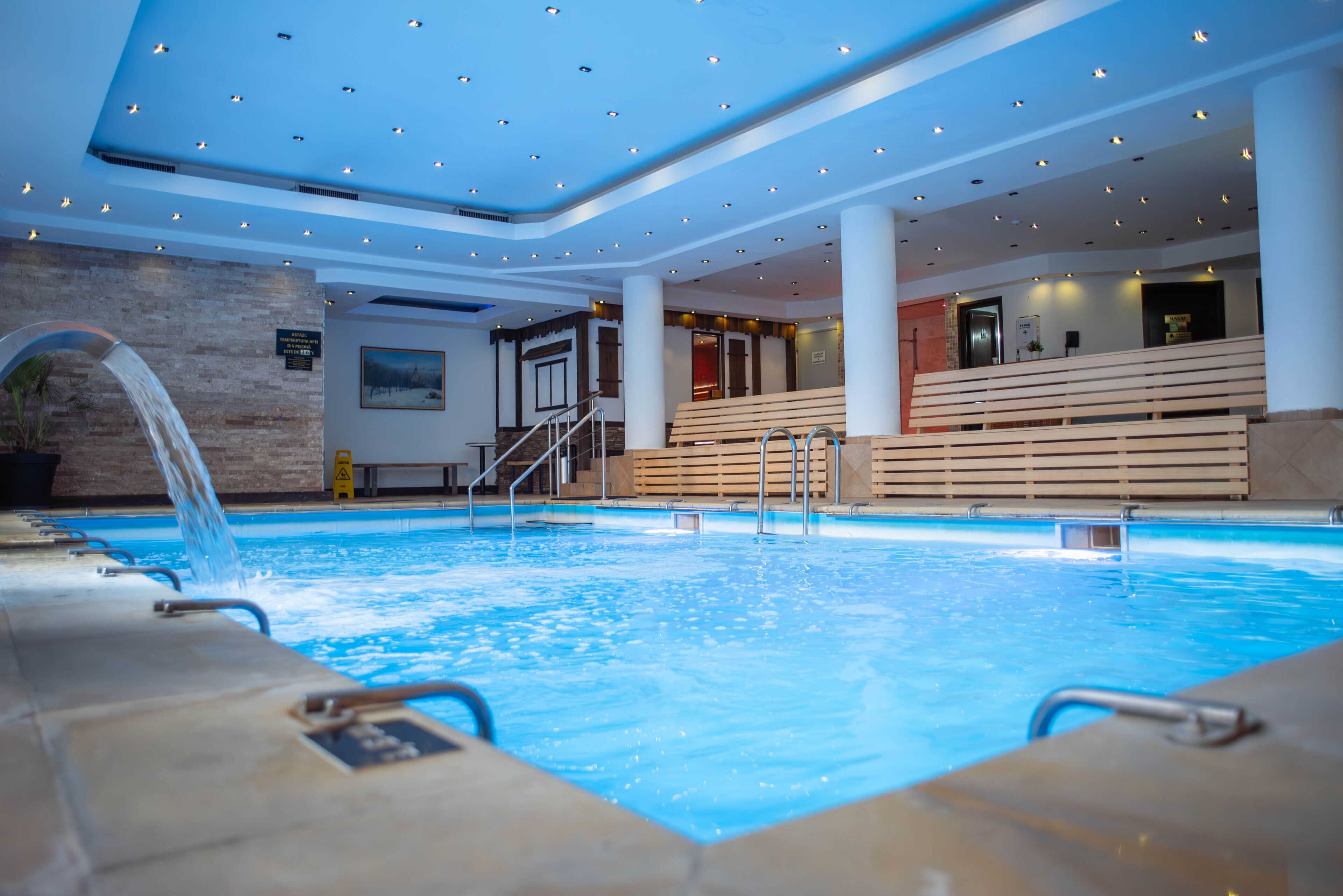 An indoor swimming pool with a waterfall feature, surrounded by wooden benches and a modern ceiling with recessed lighting.
