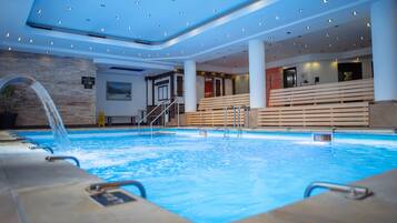 An indoor swimming pool with a waterfall feature, surrounded by wooden benches and a modern ceiling with recessed lighting.