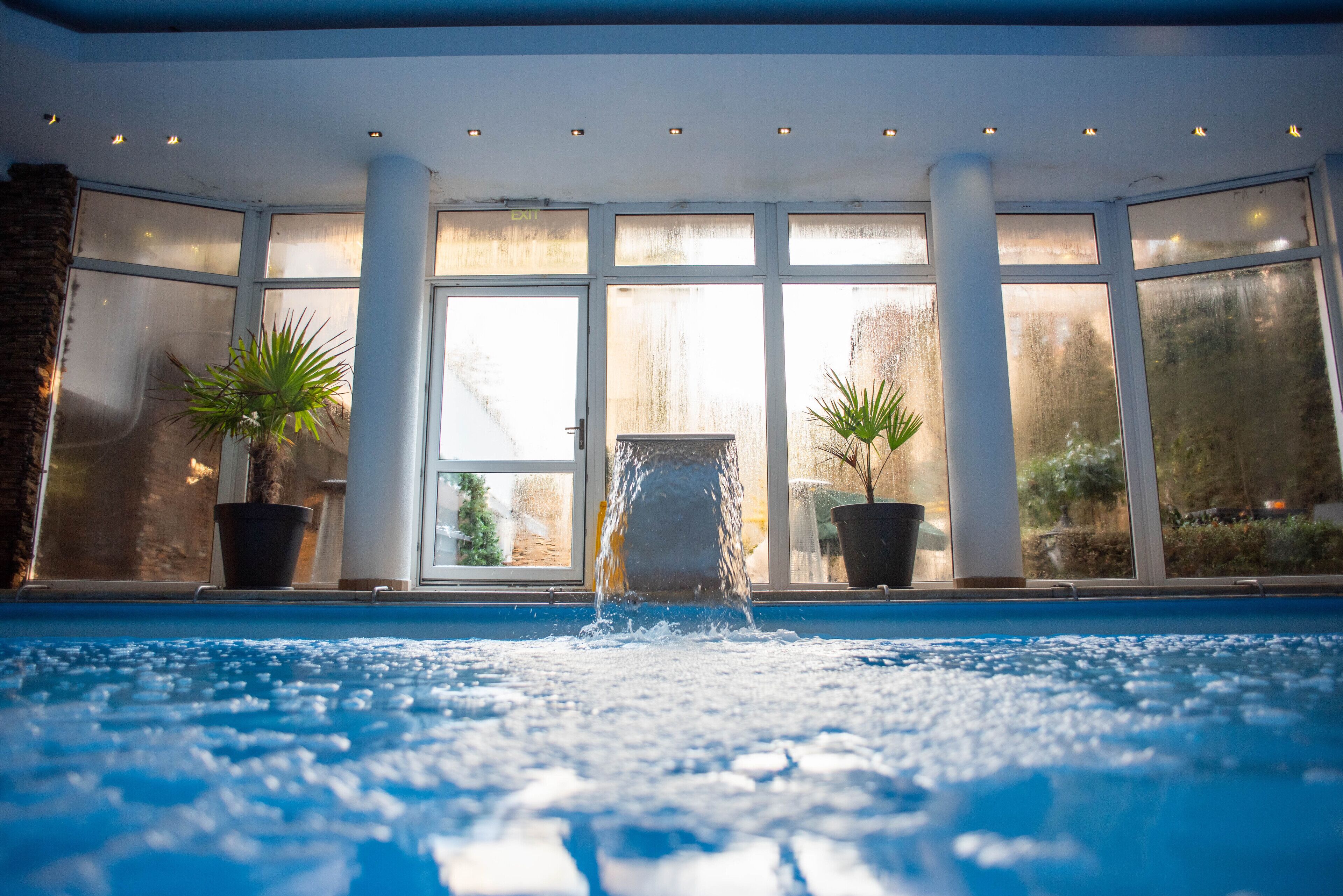 An indoor swimming pool with clear glass walls, potted plants, and a stone structure in the center.