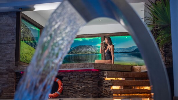 A woman standing by a pool with a waterfall feature, stone walls, and a large window with a scenic view. - Hotel Carpathia (Sinaia)