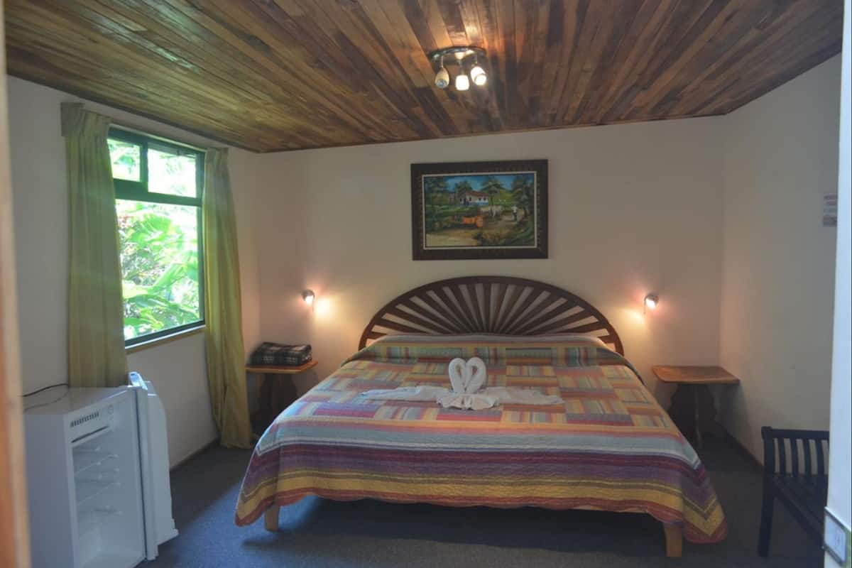 A bedroom with a wooden ceiling, a bed with a striped bedspread, a painting on the wall, and a window with curtains.