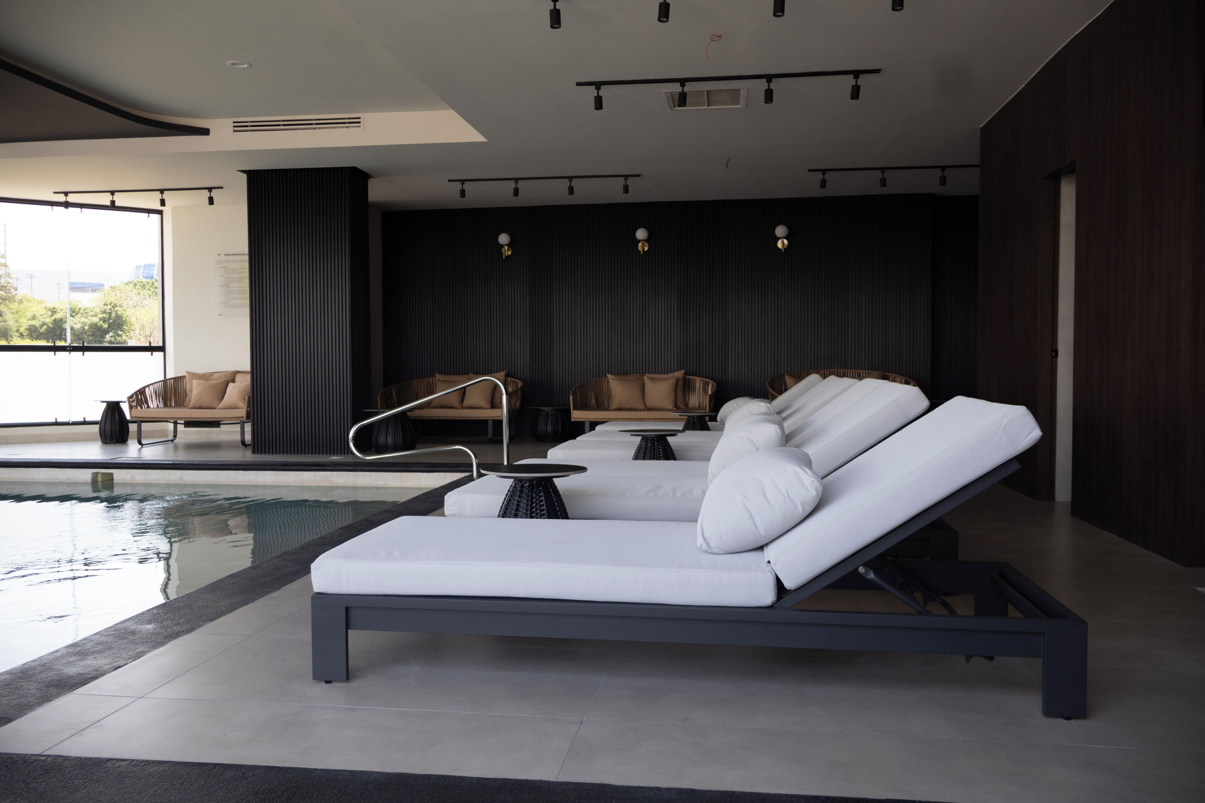 A modern indoor pool area with lounge chairs, a small table, and a view of the outdoors.