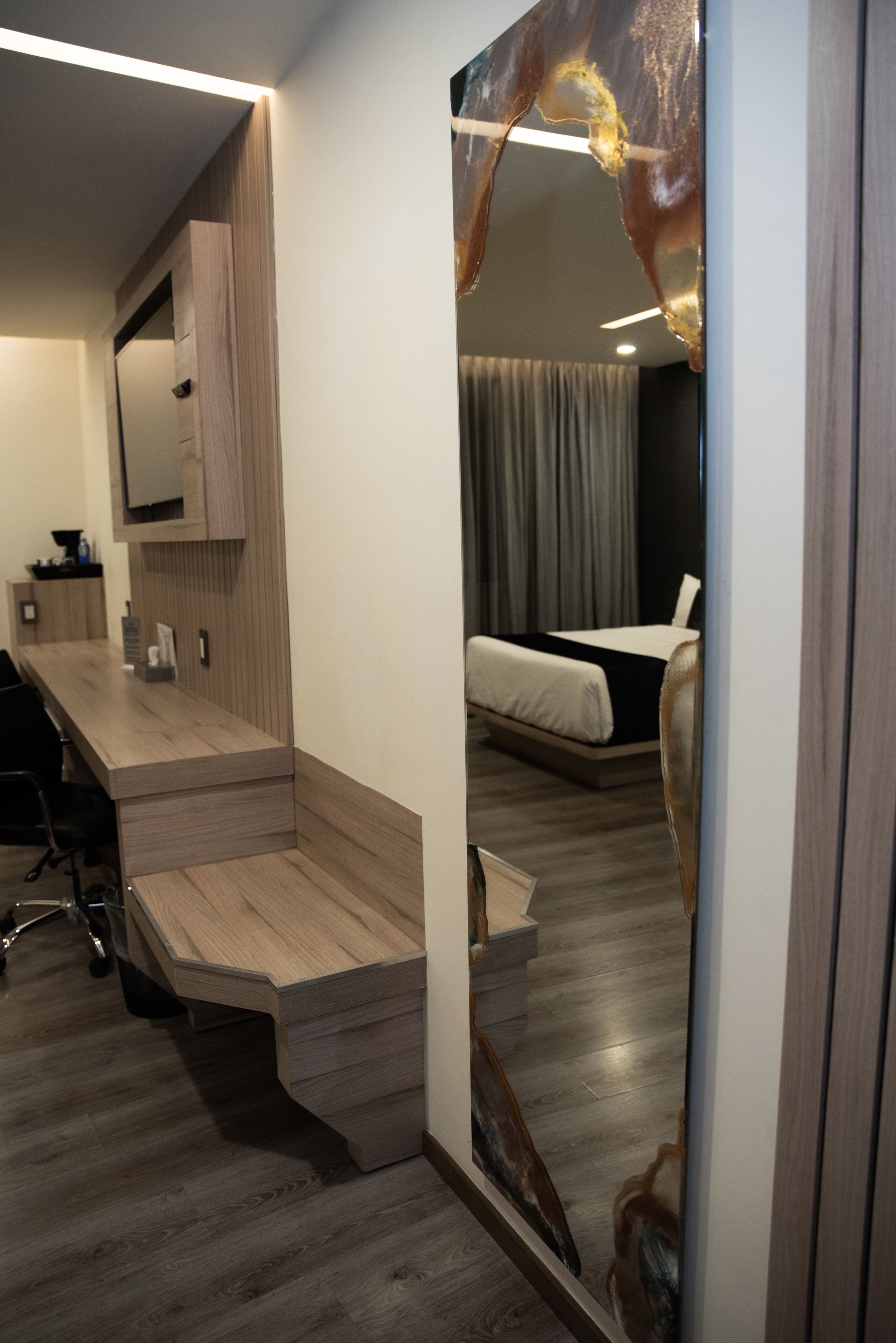 A modern hotel room with a wooden desk, a large mirror, and a bed with grey curtains.