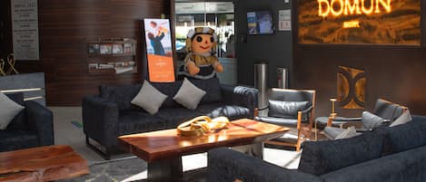A modern hotel lobby with a high ceiling, a central seating area with sofas and chairs, a wooden coffee table, and a large illuminated sign for the hotel.