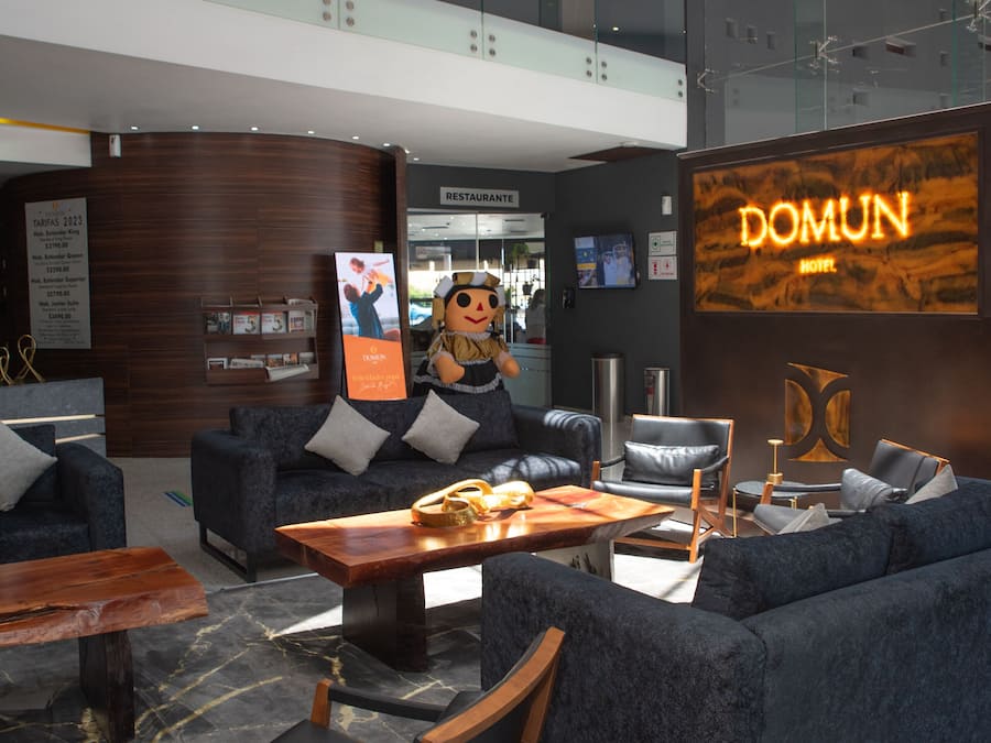 Un moderno vestíbulo de hotel con techo alto, una zona de estar central con sofás y sillones, una mesa de centro de madera y un gran letrero iluminado del hotel.
