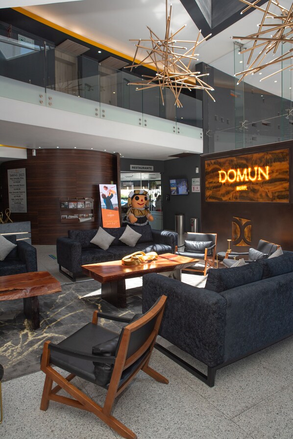 A modern hotel lobby with a high ceiling, a central seating area with sofas and chairs, a wooden coffee table, and a large illuminated sign for the hotel.