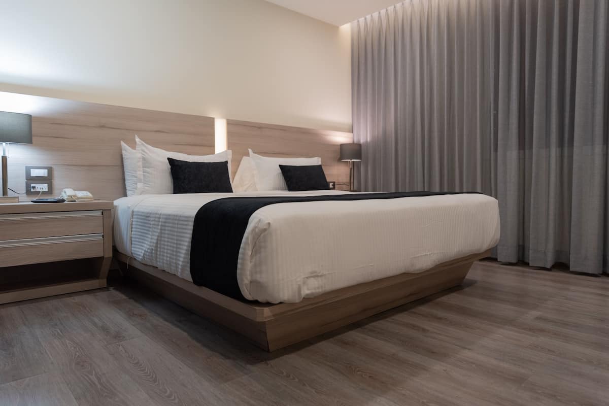 A modern hotel room with a wooden bed frame, a bedside table with a lamp, and a large window with curtains.