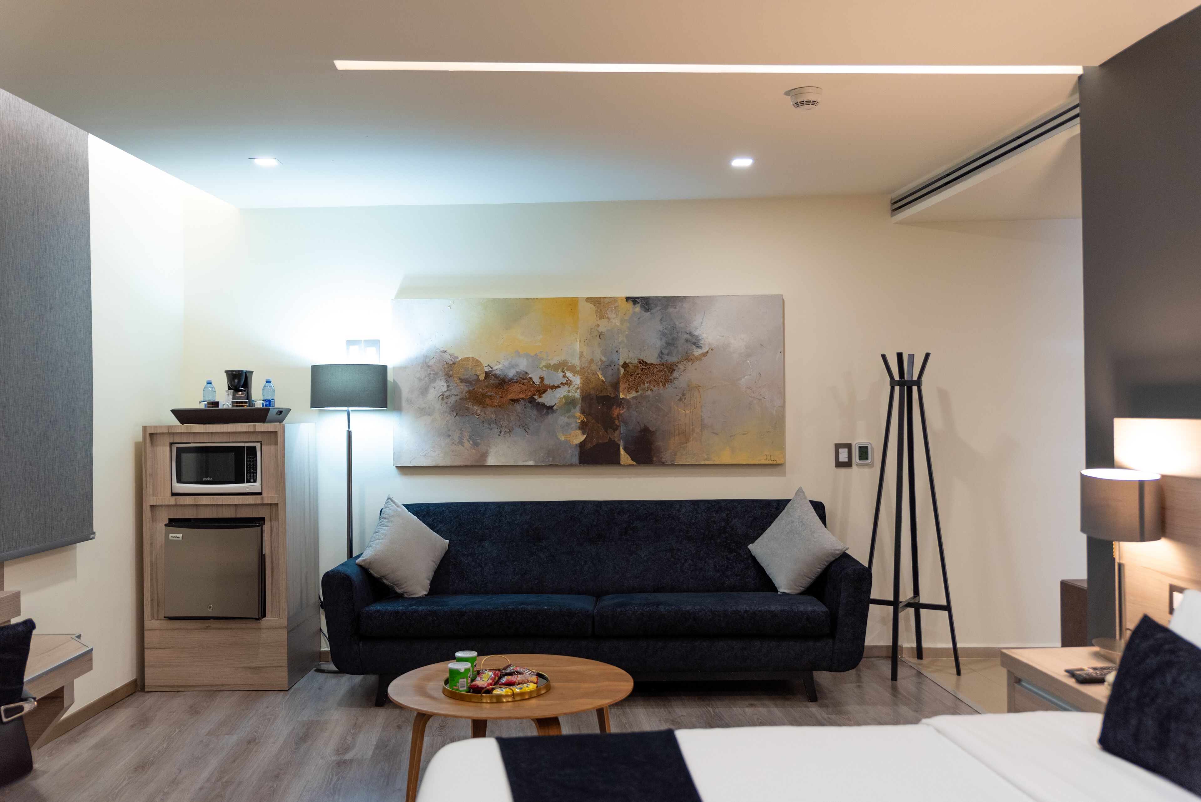 Una habitación de hotel moderna con un sofá azul oscuro, una pequeña mesa de madera con una bandeja de aperitivos, una cama con ropa blanca, un cuadro abstracto grande en la pared y una kitchenette compacta con microondas y refrigerador.