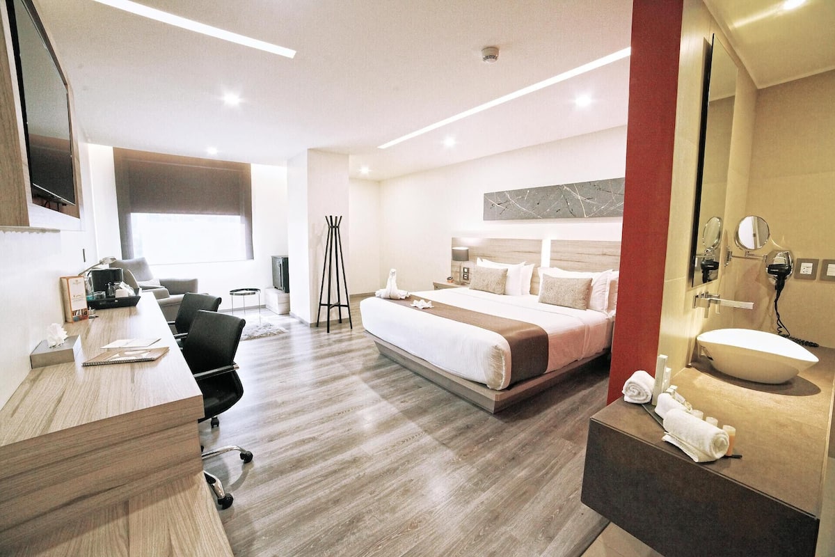A hotel room with a bed, desk, chair, TV, and a bathroom with a sink and mirror.