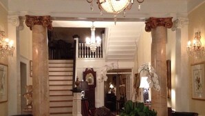 Un vaste hall d’entrée orné de colonnes en marbre, d’un escalier et d’un lustre.