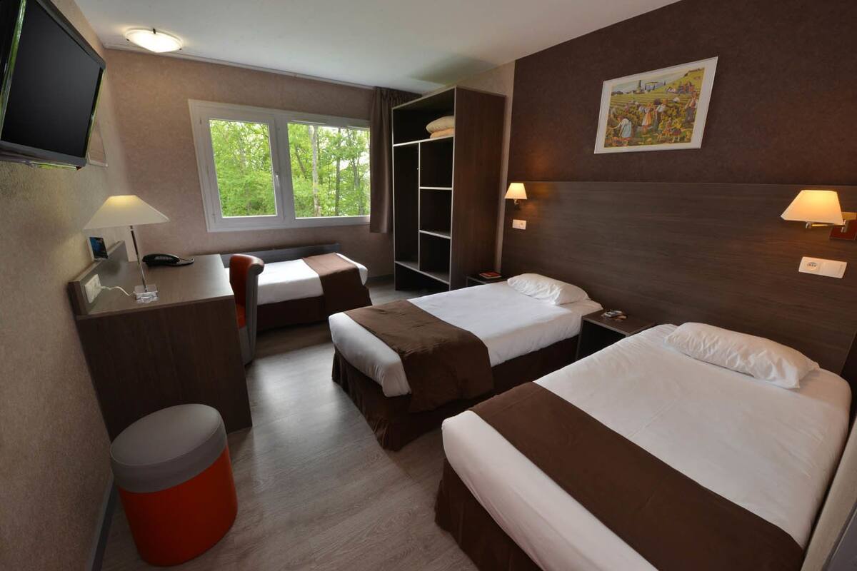 Habitación de hotel con dos camas, un escritorio, una silla, un cesto de basura y una ventana con vista a árboles.