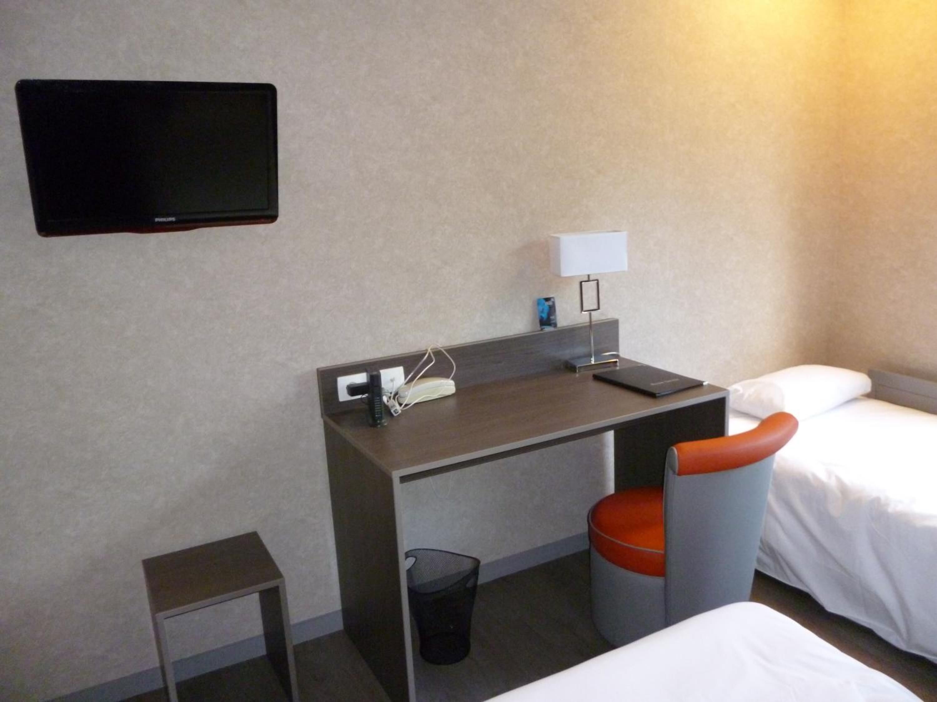 A hotel room with a desk, a chair, a TV, a lamp, and two beds.