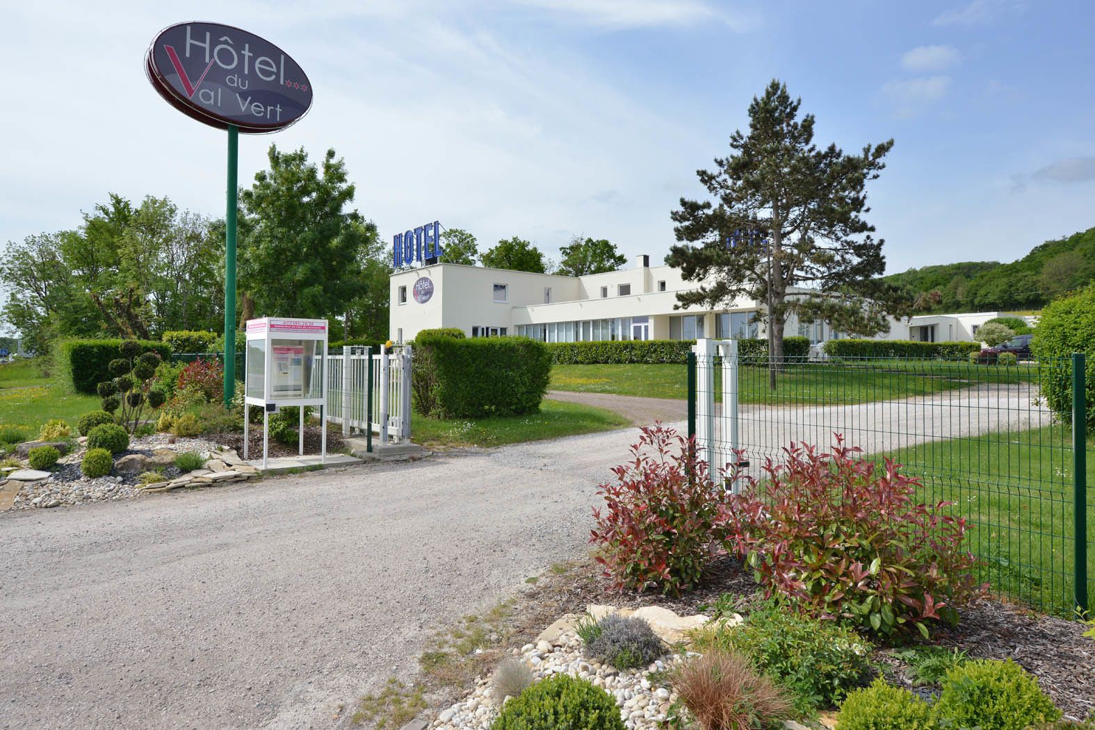 En hotelindgang med et skilt, en grusvej og et indhegnet område med grøn bevoksning.