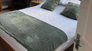Quarto com claraboia, cama com roupa de cama verde, mesa de cabeceira, abajur e uma cadeira.
