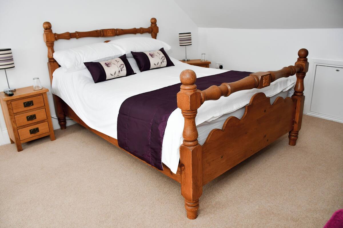 Una cama con dosel de madera, ropa blanca y detalles morados, una mesita de noche con lámpara y un cajón pequeño.