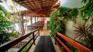 A covered outdoor dining area with wooden tables and chairs, surrounded by greenery and a stone pathway.