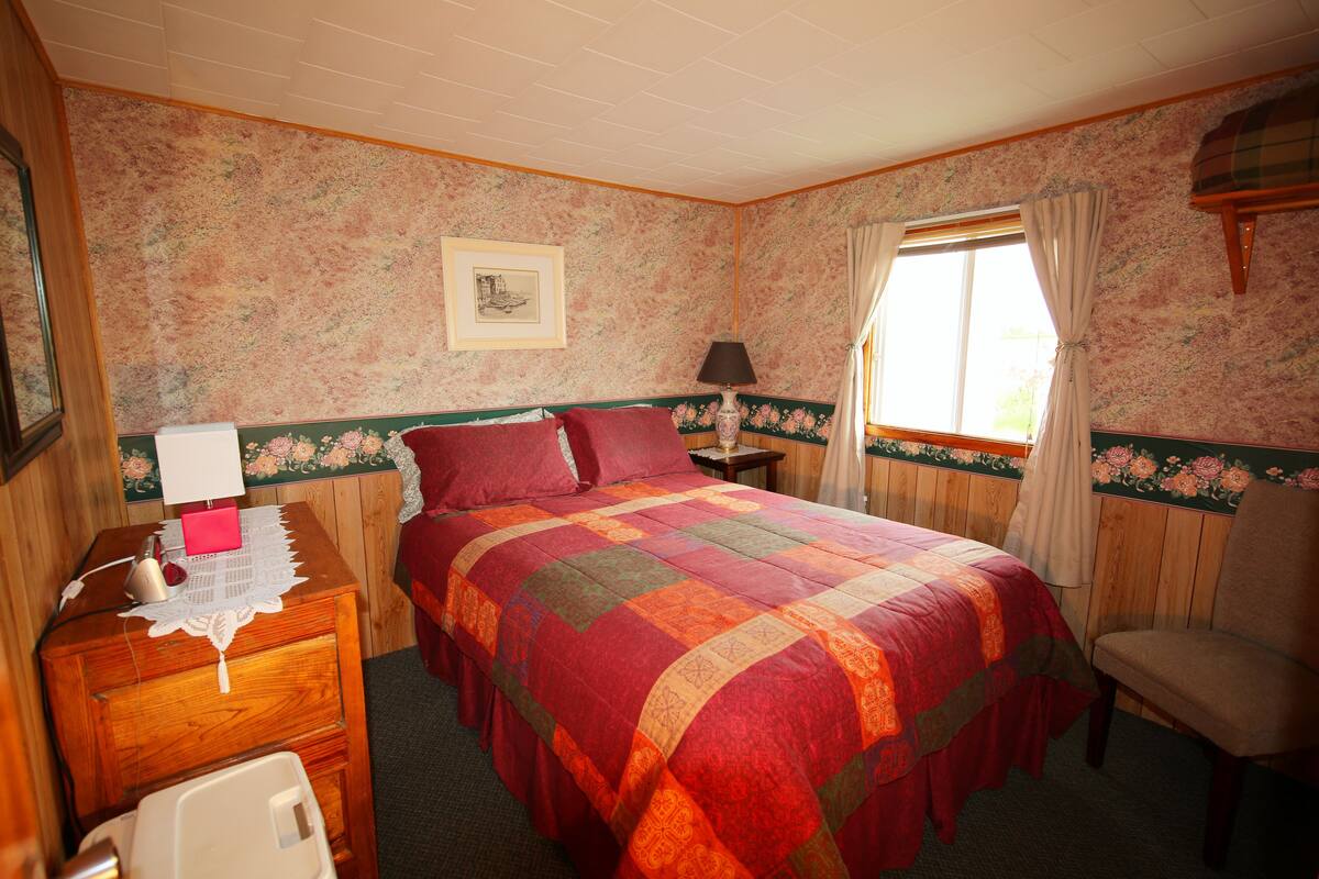 A bedroom with a bed, a chair, a nightstand, a lamp, and a window with curtains.