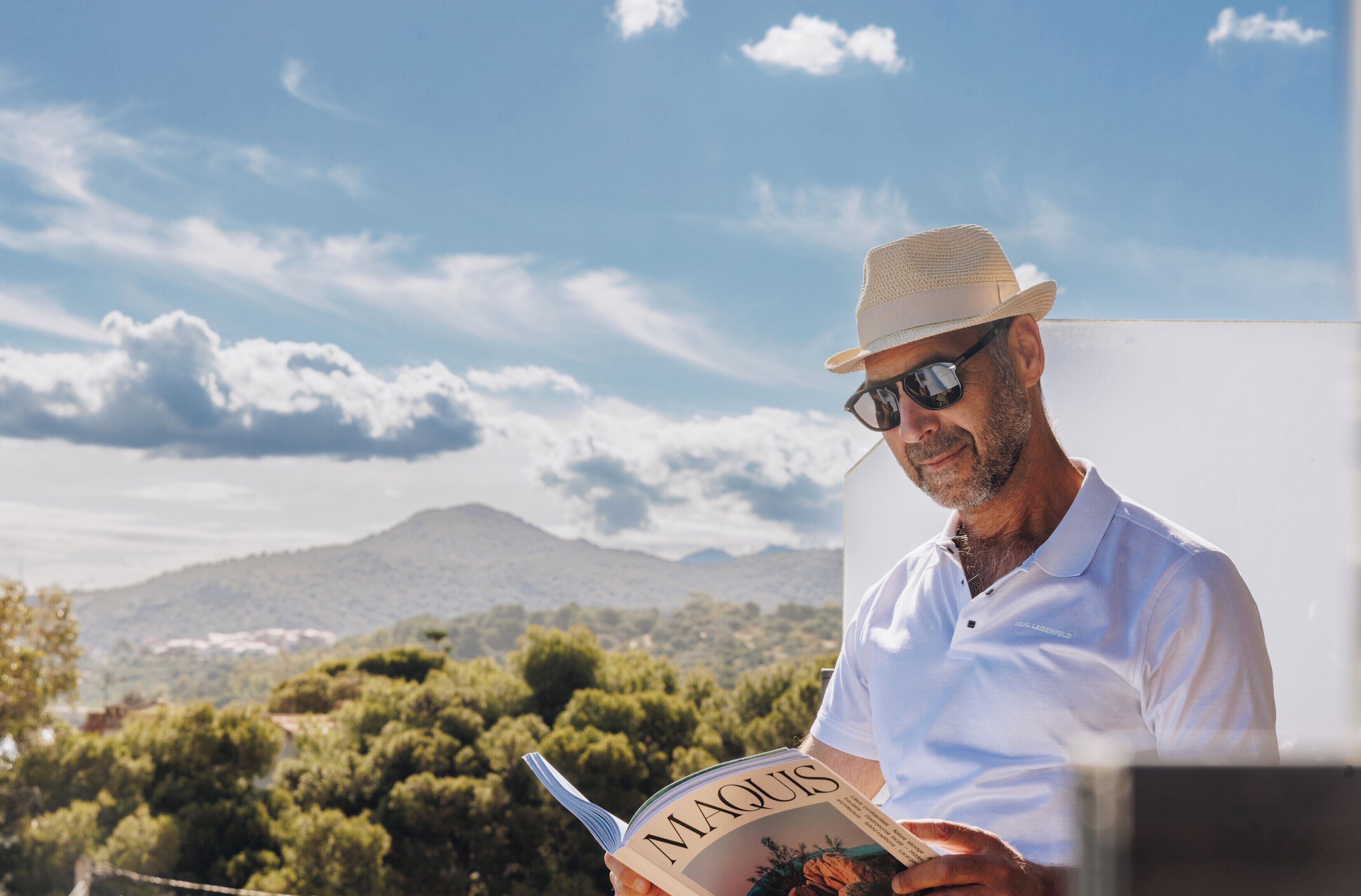 A person reading a book titled "MAQUIS" outdoors with a mountainous landscape in the background.