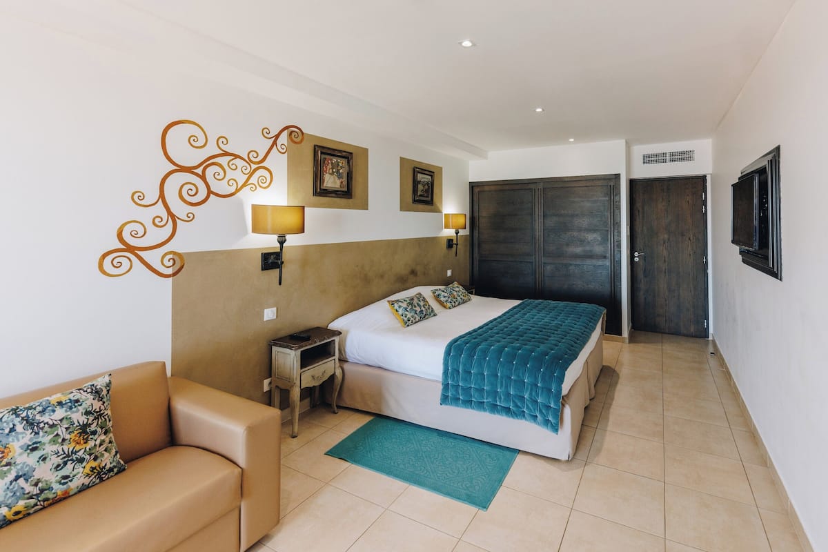 A hotel room with a bed, a nightstand, a sofa, a wall-mounted TV, and a door.
