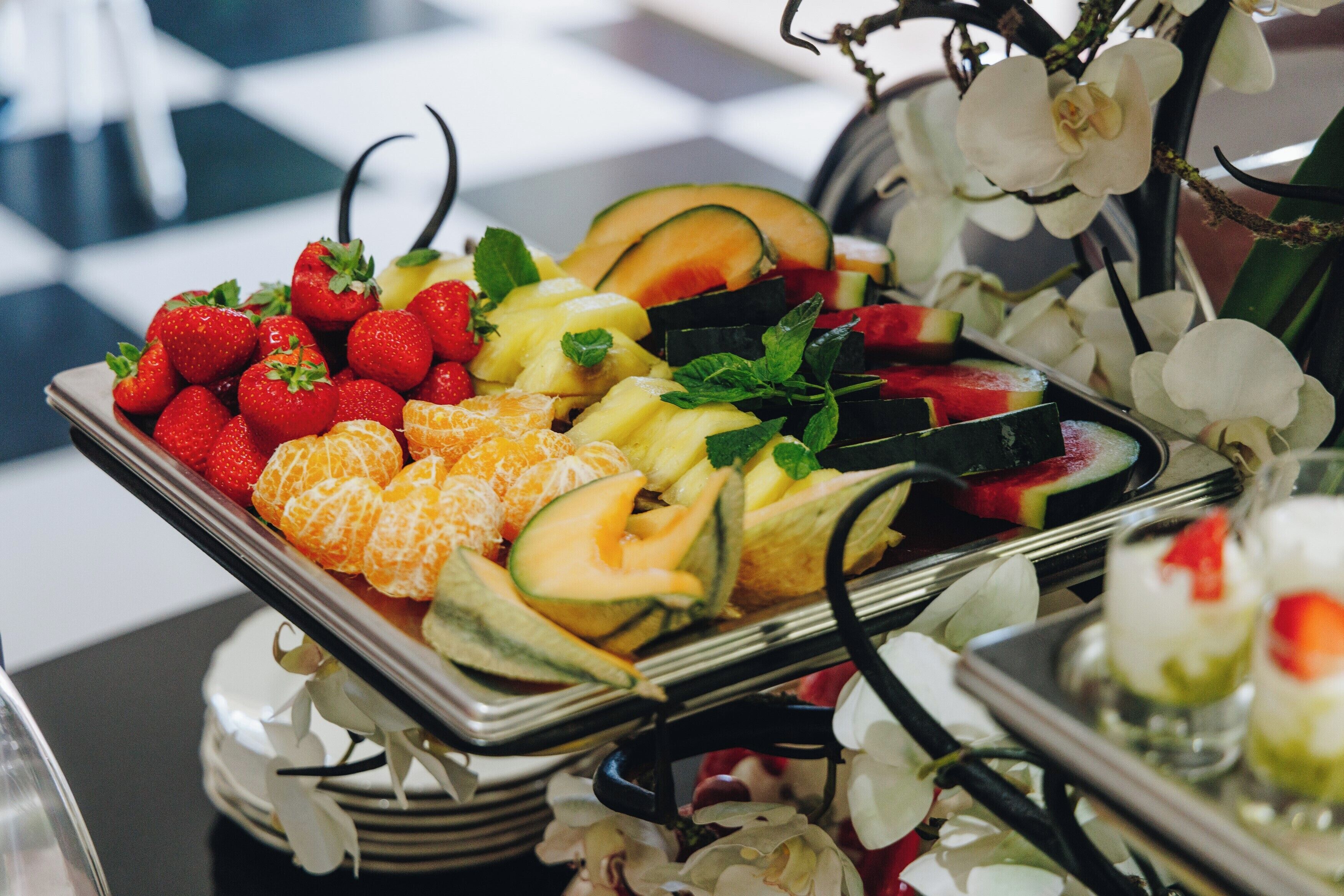 Una bandeja con frutas variadas, como fresas, naranjas y trozos de melón, dispuestas sobre una bandeja metálica con una orquídea decorativa al fondo.