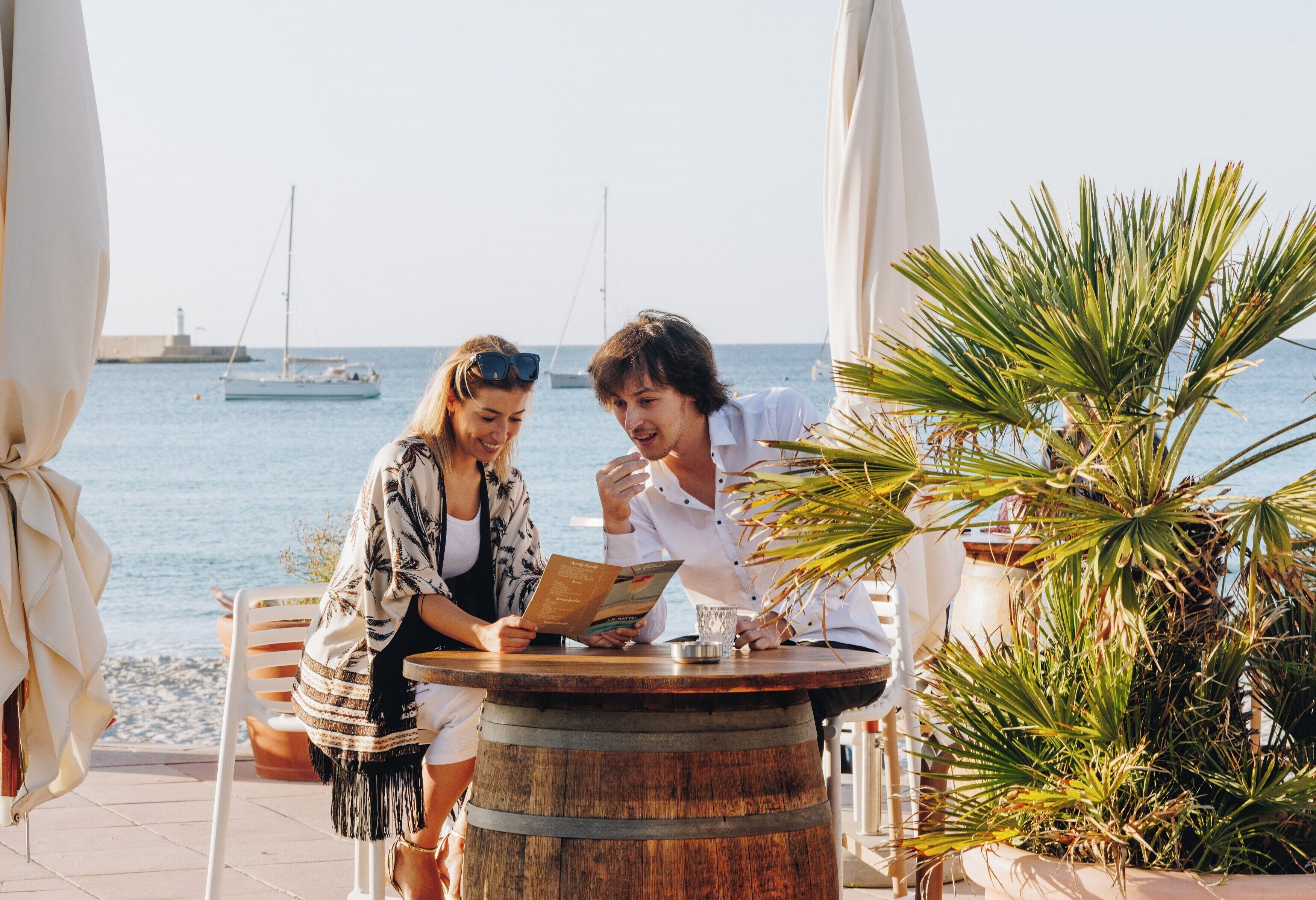 Twee personen zitten aan een houten tafel met uitzicht op zee en boten op de achtergrond.