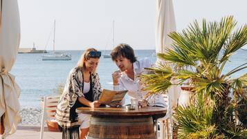 Twee personen zitten aan een houten tafel met uitzicht op zee en boten op de achtergrond.
