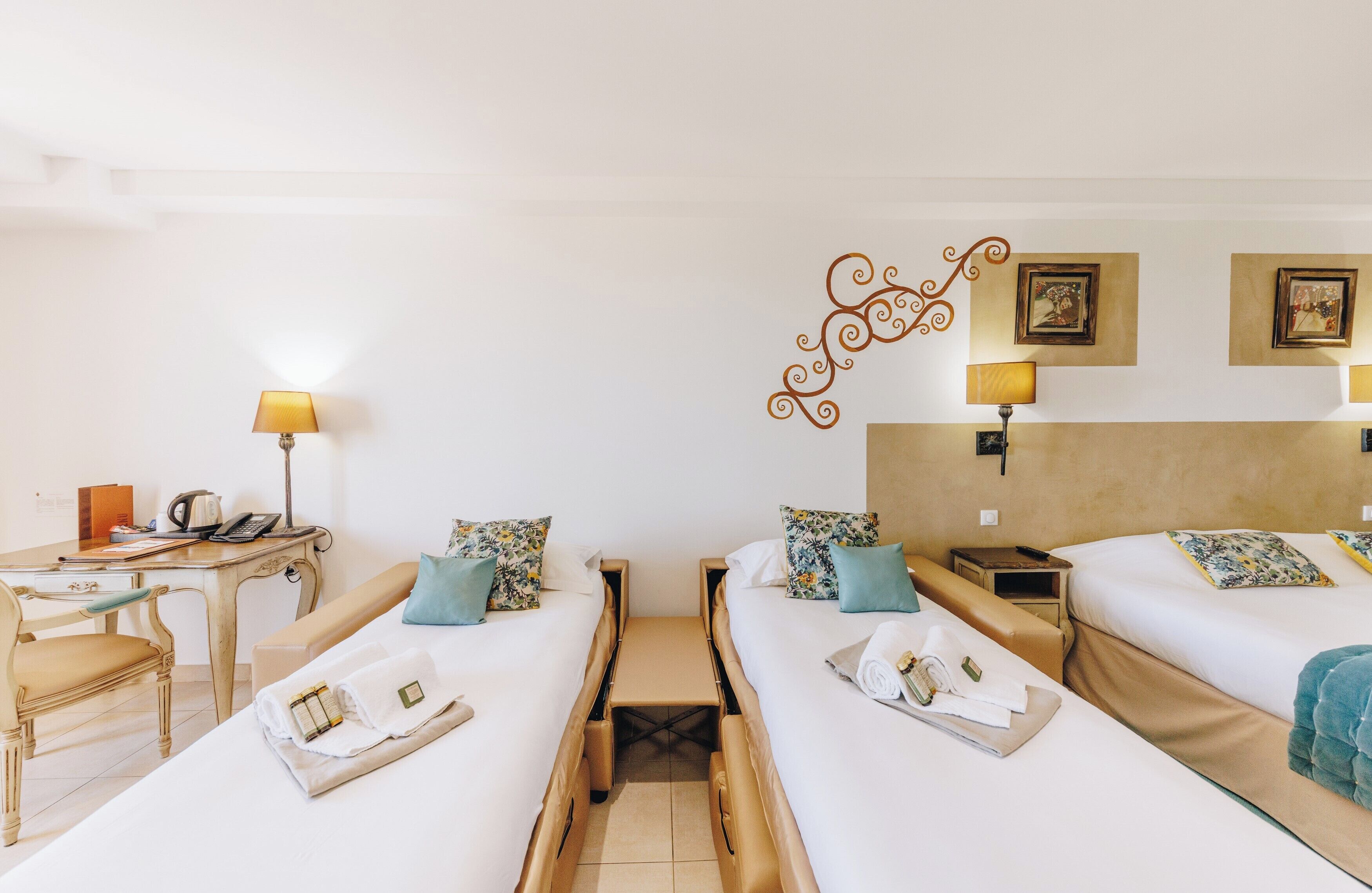 Habitación de hotel con dos camas individuales, un escritorio con lámpara, una silla y una mesita con hervidor.