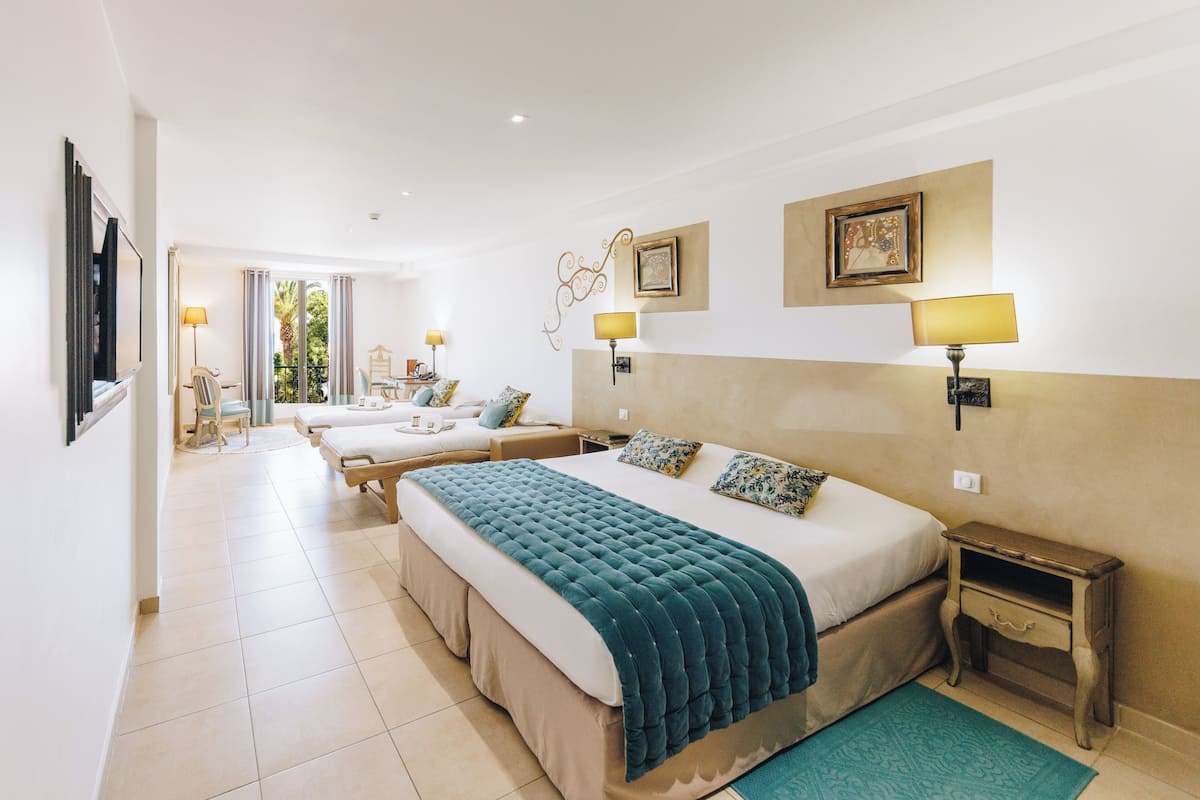 A spacious hotel room with a large bed, a bedside table, a small desk, and a chair. There is a window with curtains, a wall-mounted TV, and a framed picture on the wall.