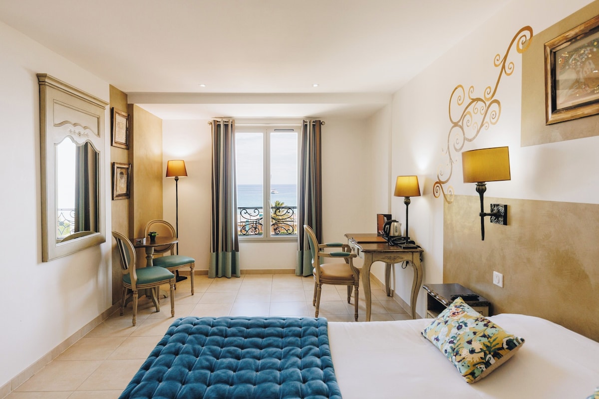 A hotel room with a bed, two chairs, a desk, a lamp, and a view of the sea through a window.