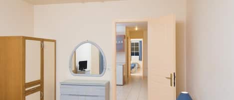 Un dormitorio con un armario de madera, un aparador con cajones, un espejo, una mesita de noche con lámpara y una puerta que da a un pasillo.
