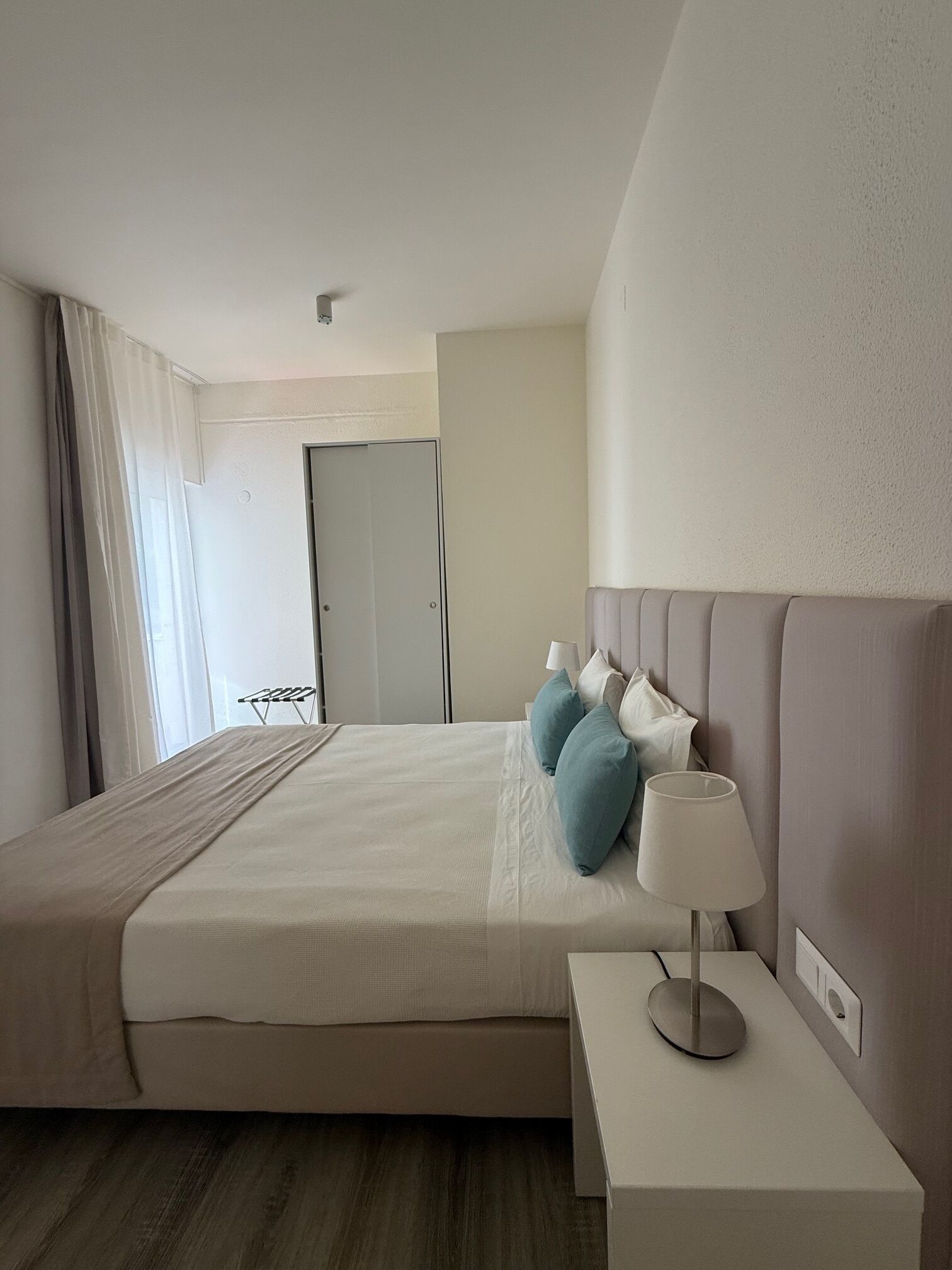 A hotel room with a bed, bedside table, lamp, and a view of a hallway with a door.